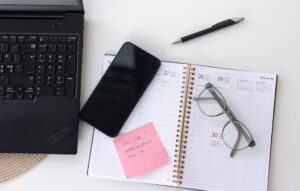 Pöydällä kalenteri, puhelin, läppäri ja silmälasit.